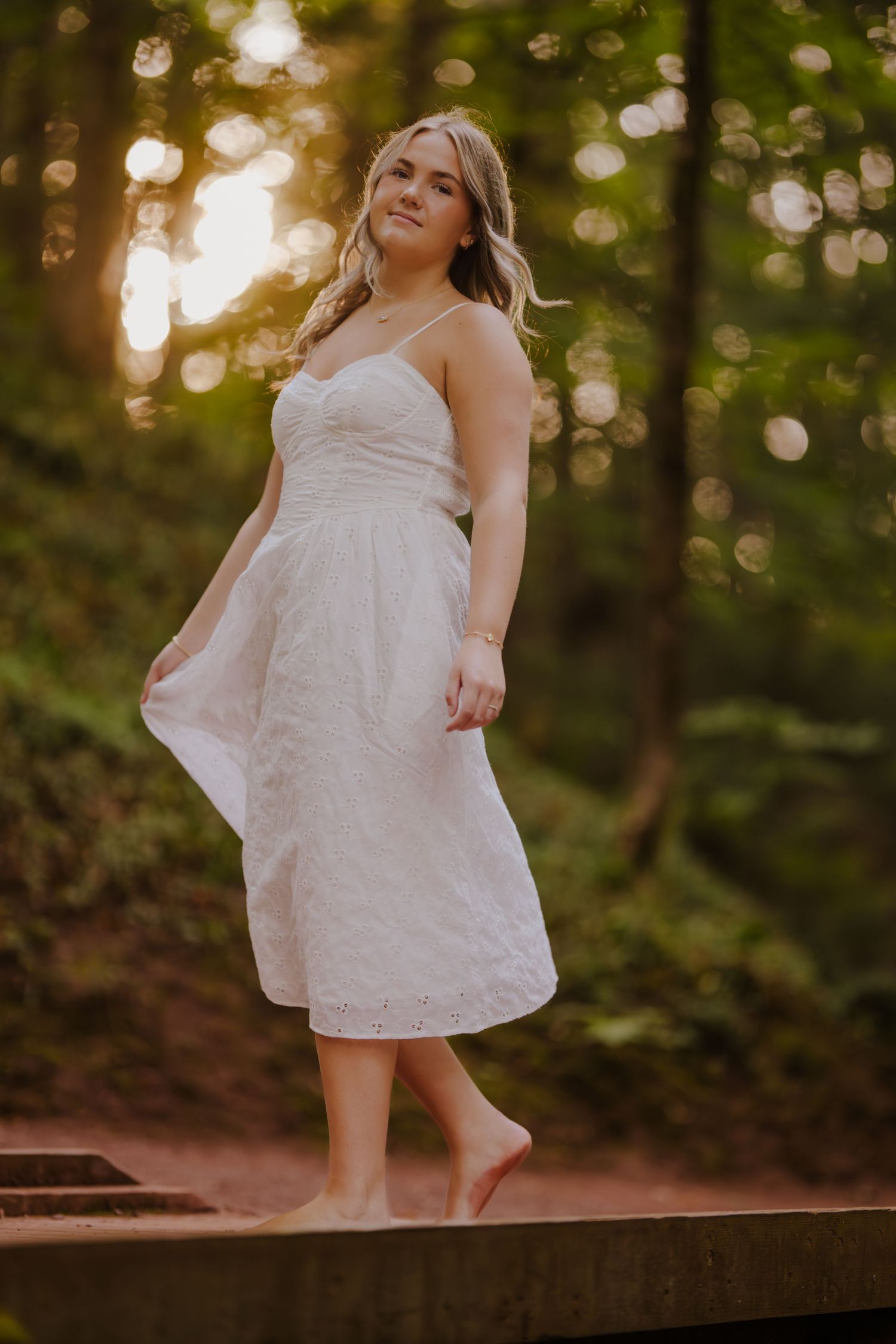 Woman in white dress stands barefoot on concrete ledge at sunset in wooded setting.