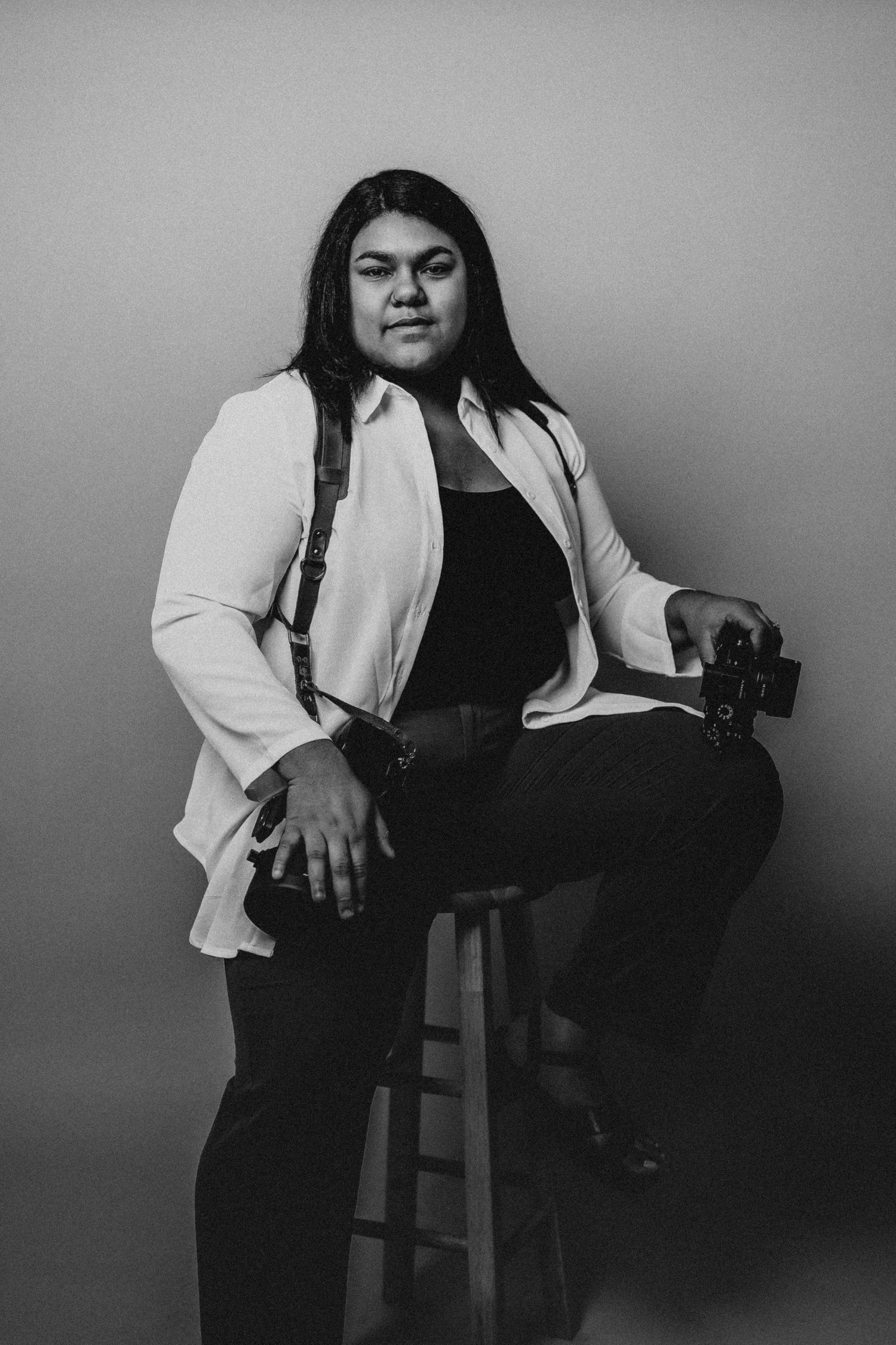 A black and white portrait of a photographer holding camera equipment while seated on a stool.