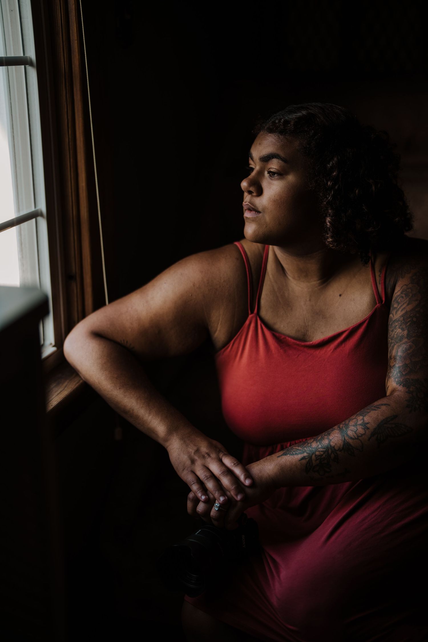 Figure in red tank top gazing thoughtfully by window in moody lighting.