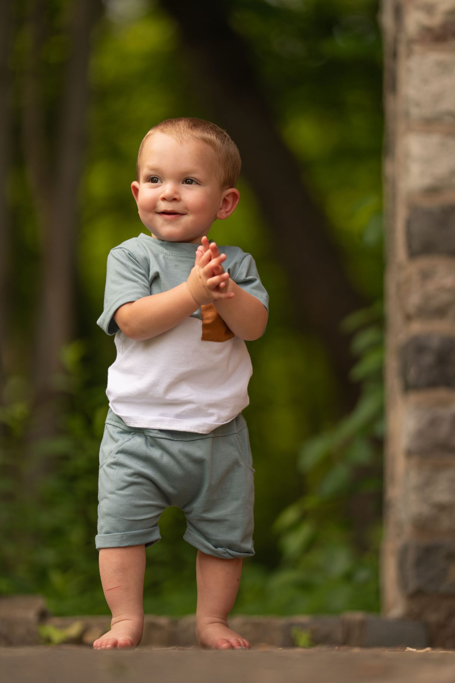 Baby wearing light blue outfit standing outdoors against blurred natural background.