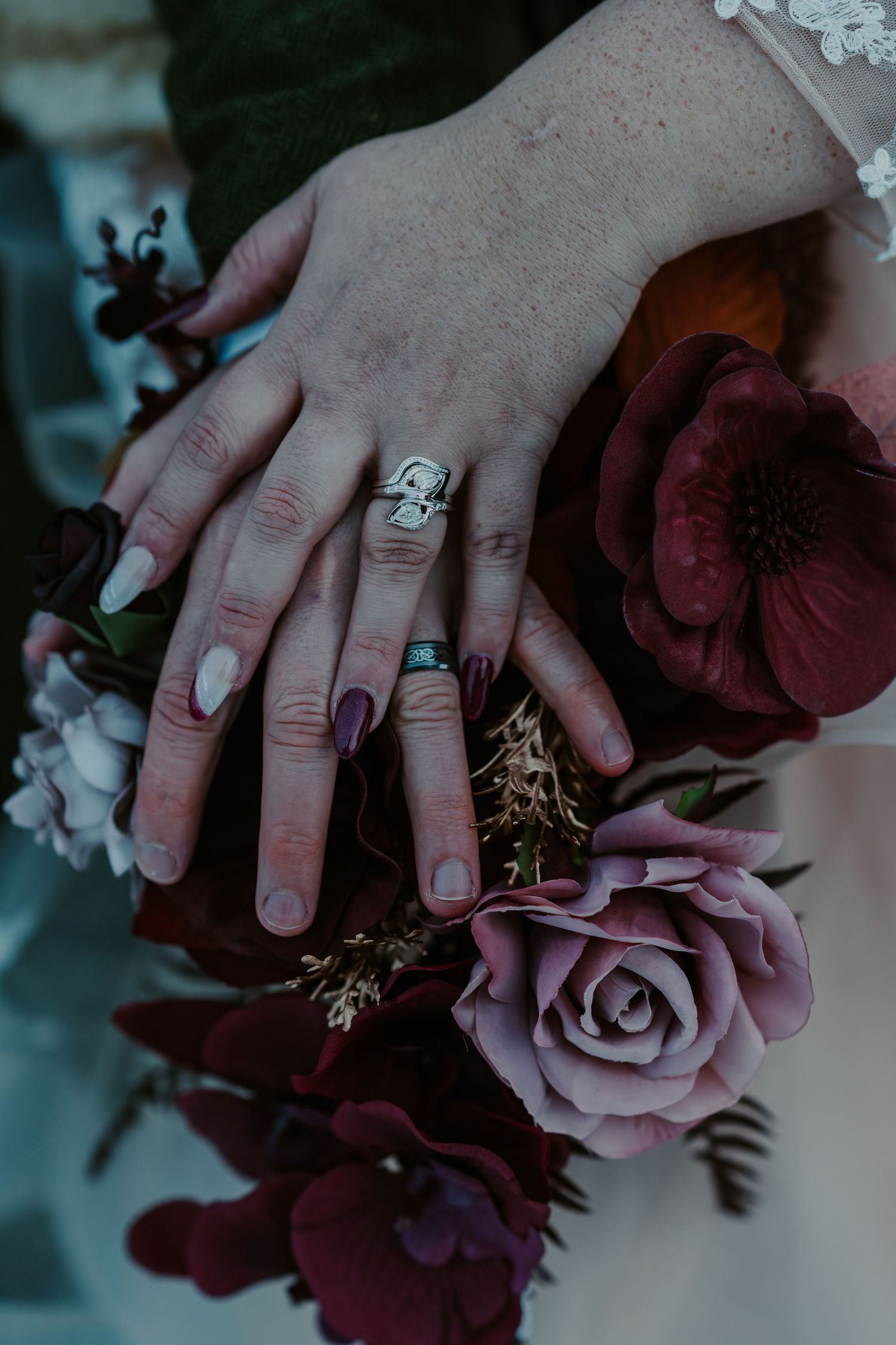 Close-up details of hands with rings and burgundy roses in a wedding bouquet.