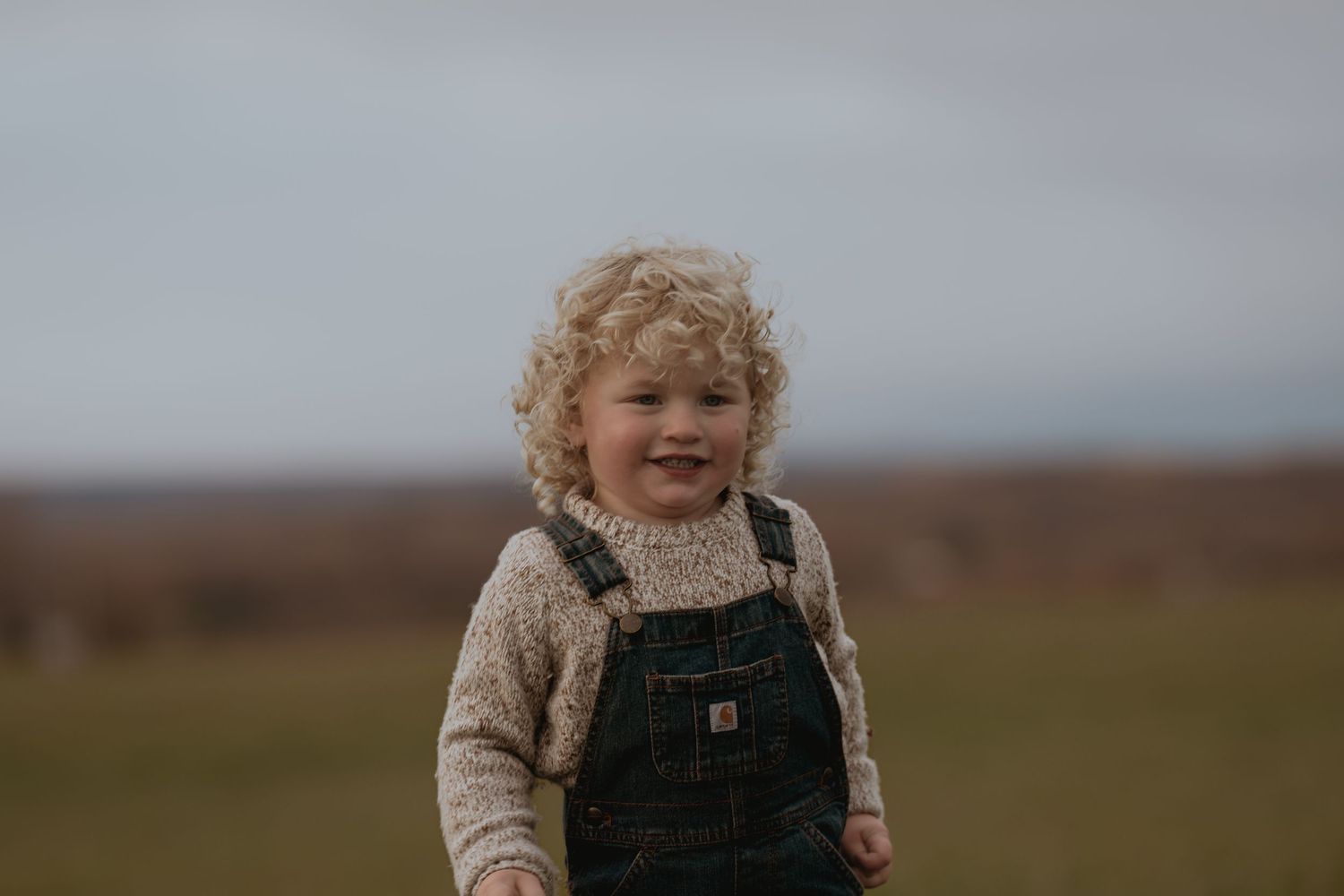 Curly-haired toddler wearing overalls shows pure joy while playing outdoors.