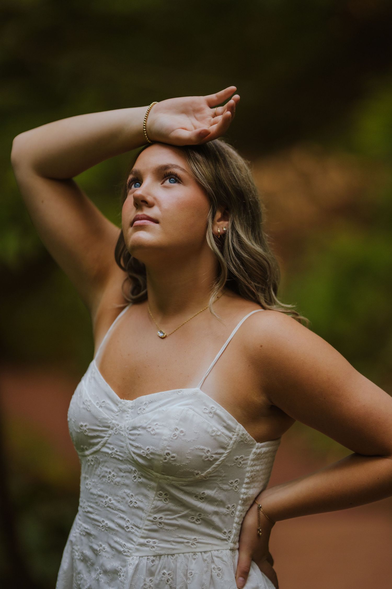 Woman in a white embroidered dress poses with arm raised against a blurred nature background.