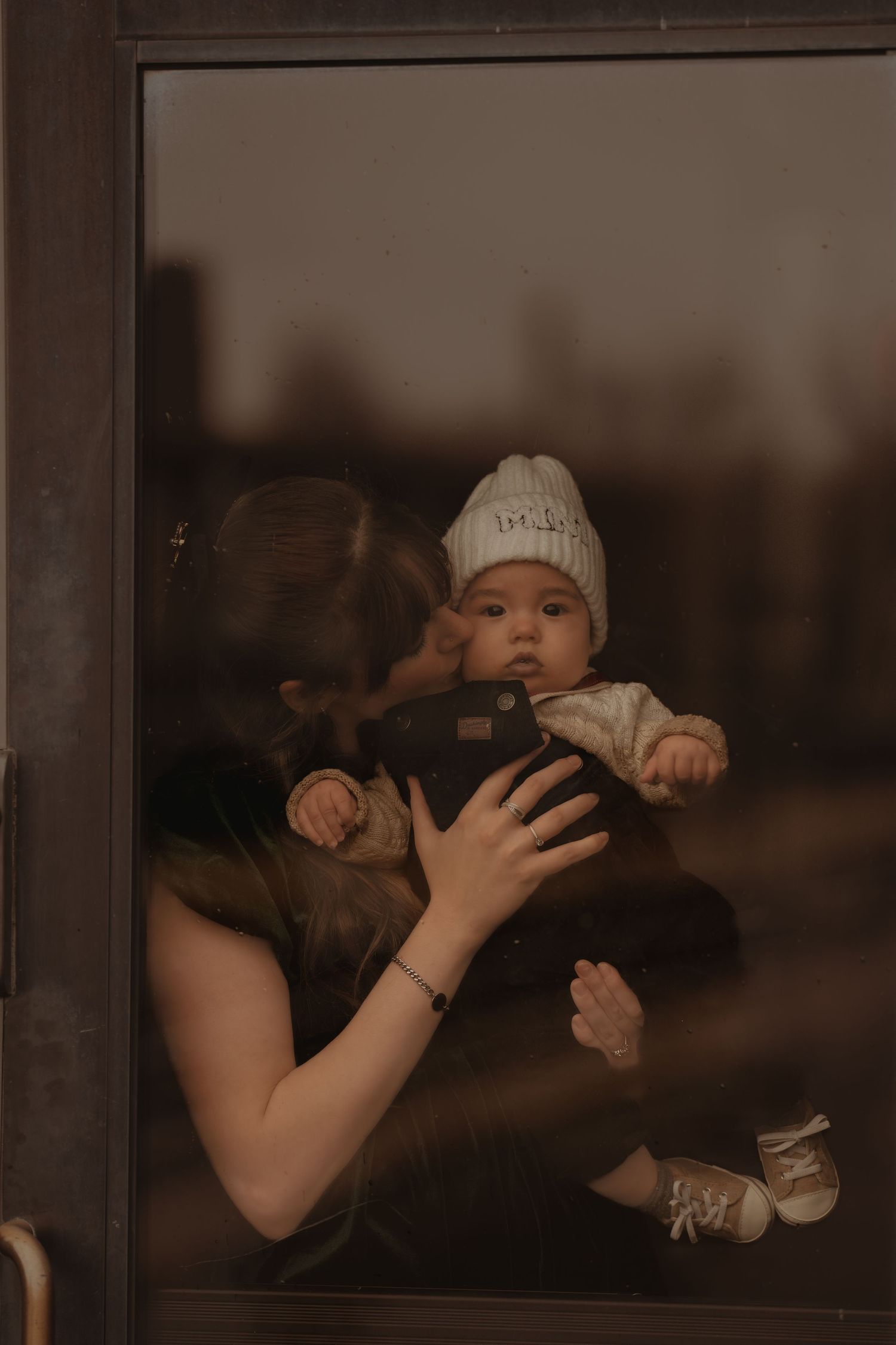 Parent holding baby in white knit hat seen through window reflection.