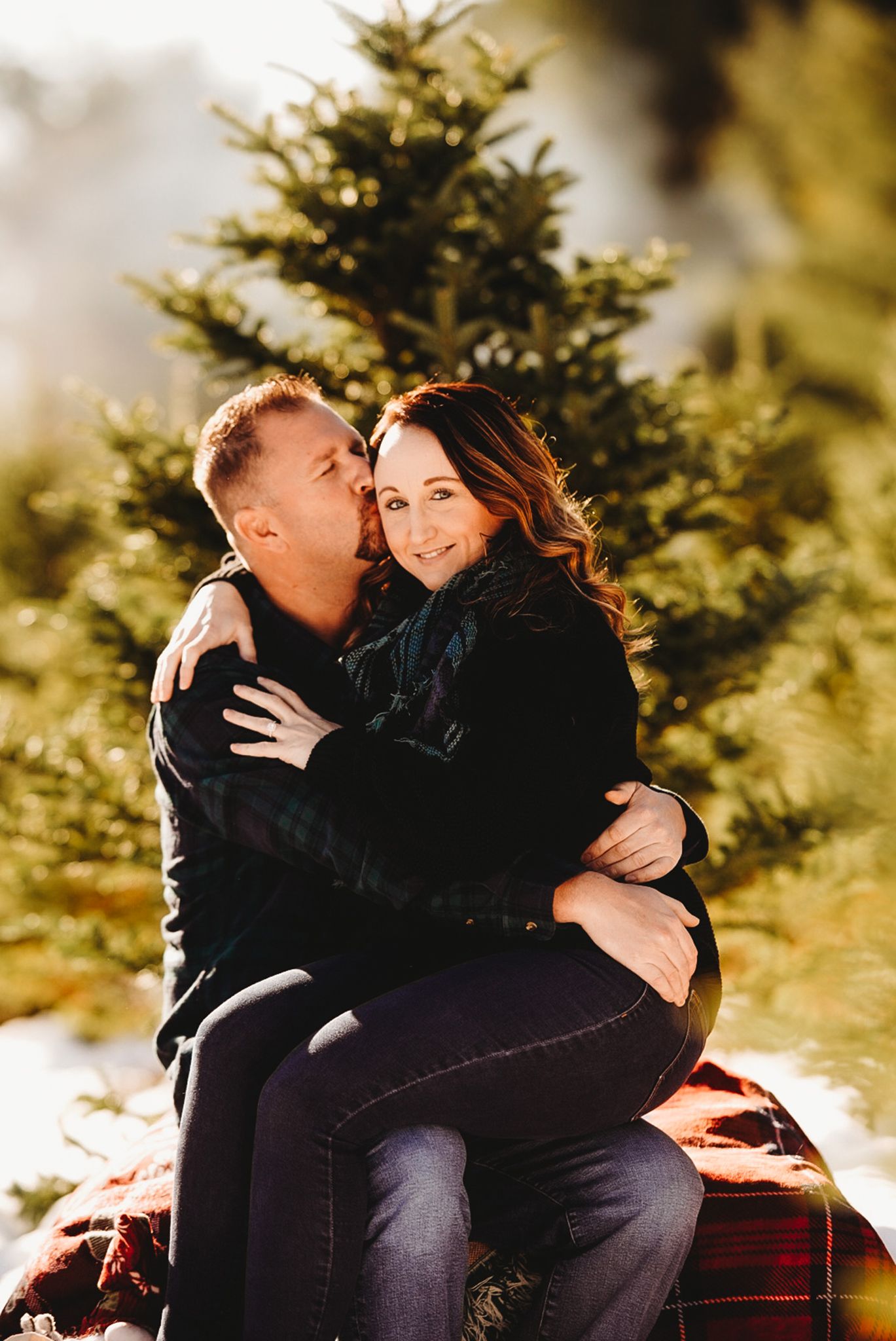 A cozy winter photoshoot near evergreen trees shows an intimate couple embracing.