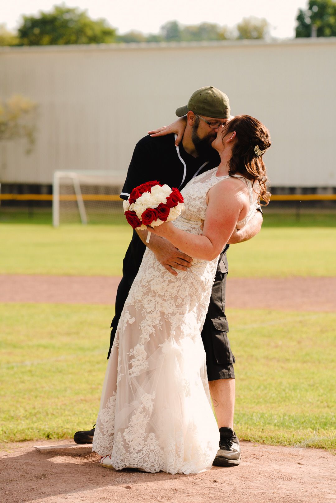 A couple shares an intimate dance dip pose on a baseball diamond during what appears to be a wedding photoshoot.