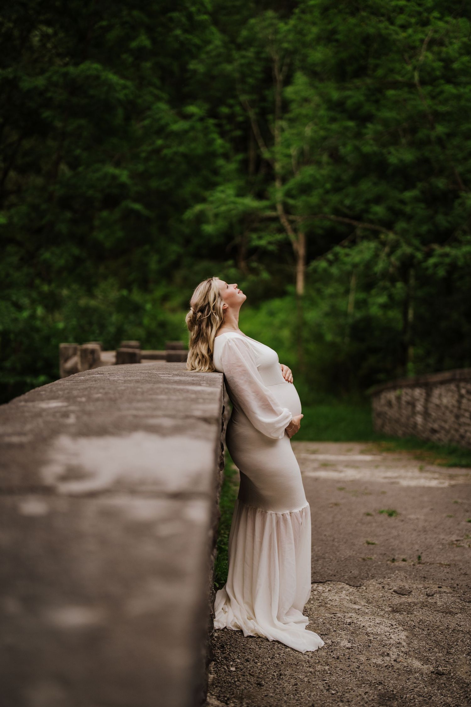 A maternity photo on a stone bridge features a flowing cream dress against vibrant forest greenery.