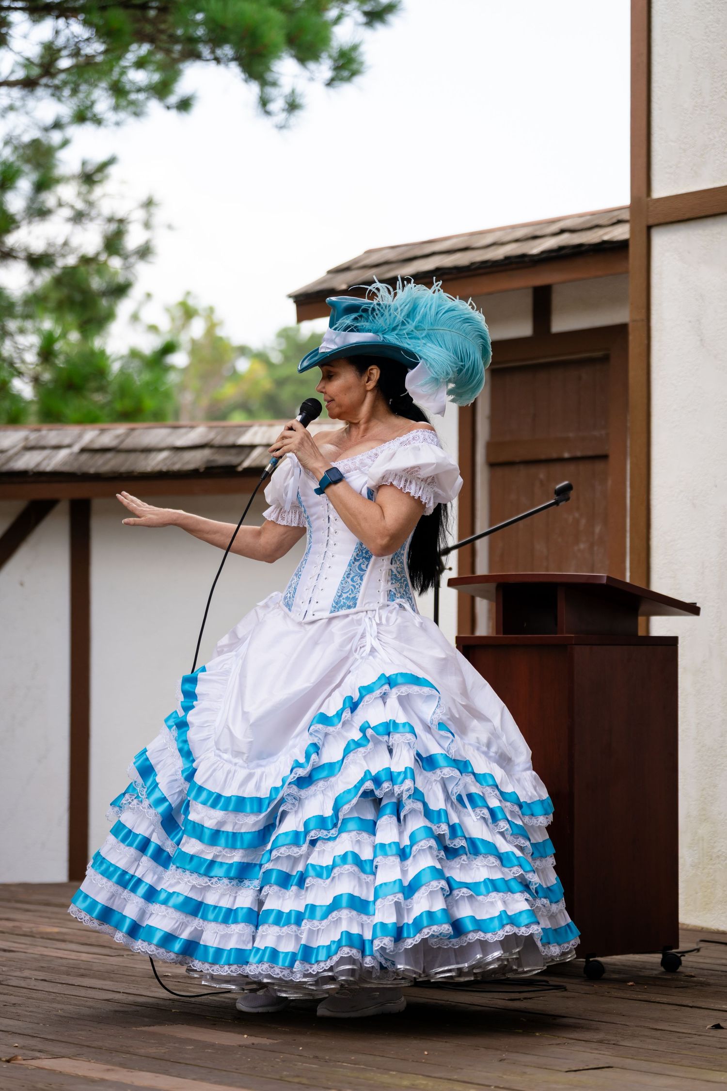 Woman in white and blue Victorian dress with feathered hat singing into microphone on outdoor stage.