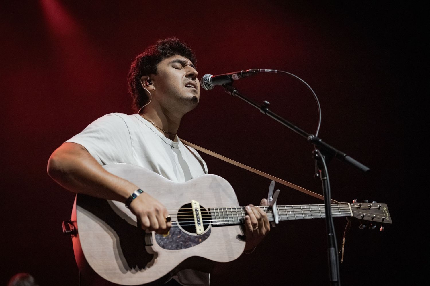 Young man in white t-shirt playing acoustic guitar and singing into microphone on stage under red lighting.