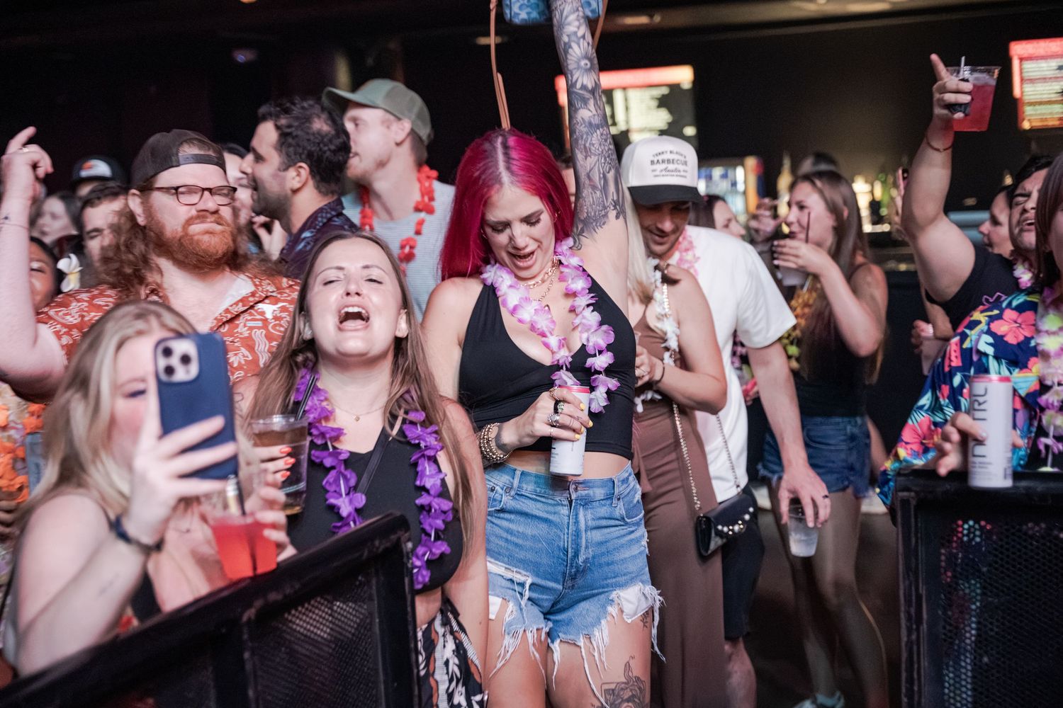 Crowd of people enjoying a lively bar event, with a woman in denim shorts and lei laughing joyfully.