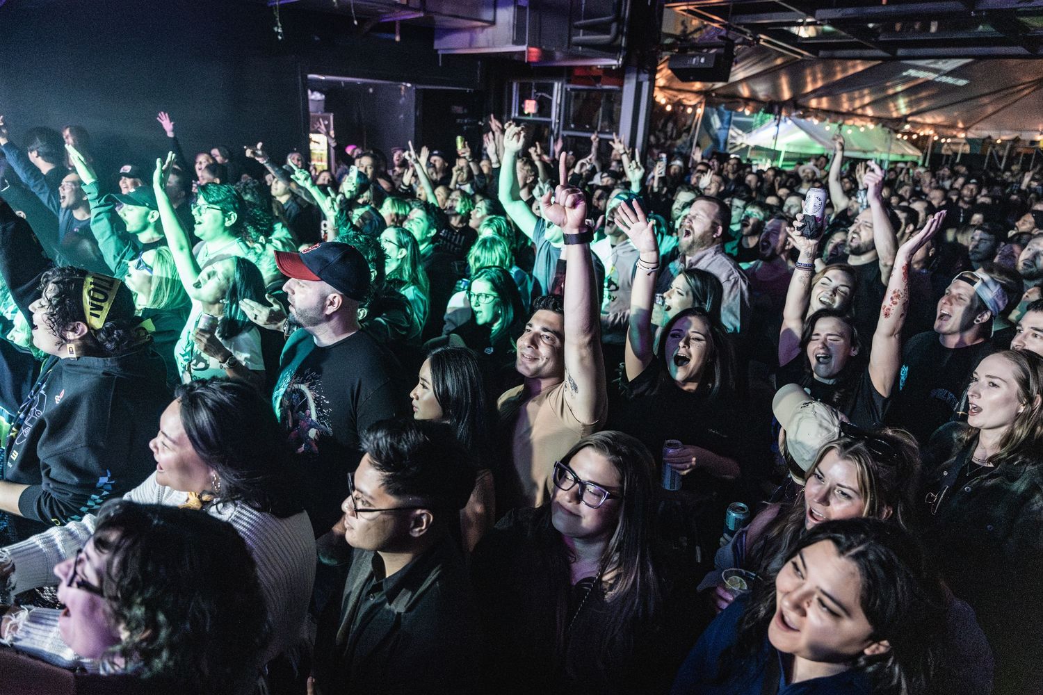 Concert crowd raises hands at an indoor music venue illuminated by green lighting and stage effects during live performance.