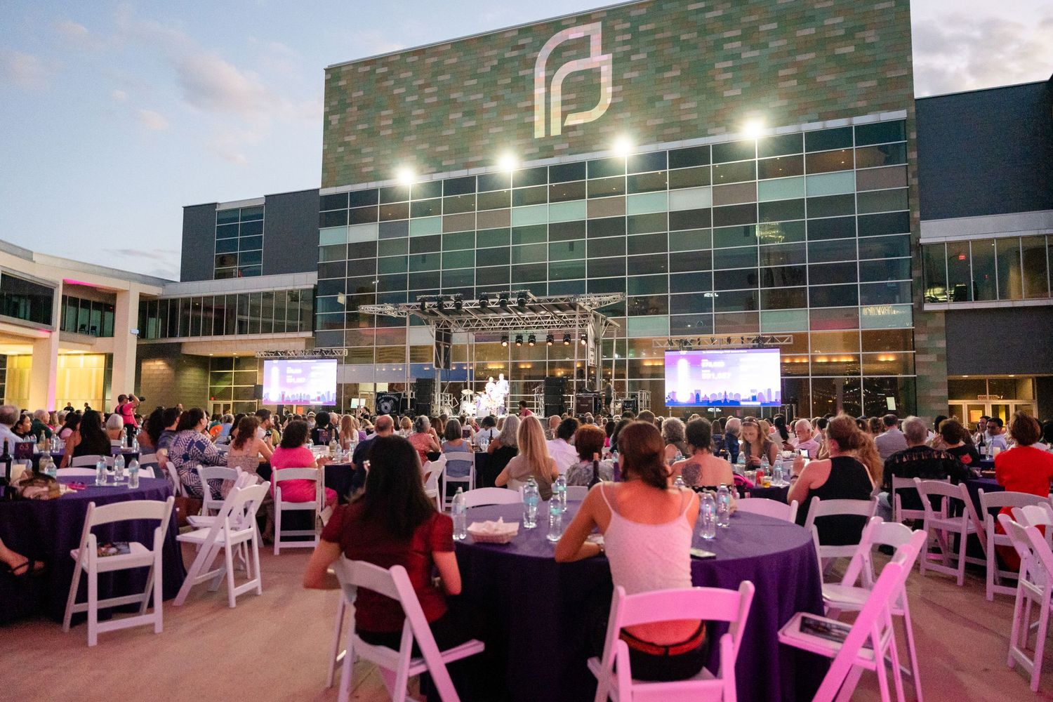 Outdoor evening event setup with white chairs and tables in front of modern building with illuminated logo.