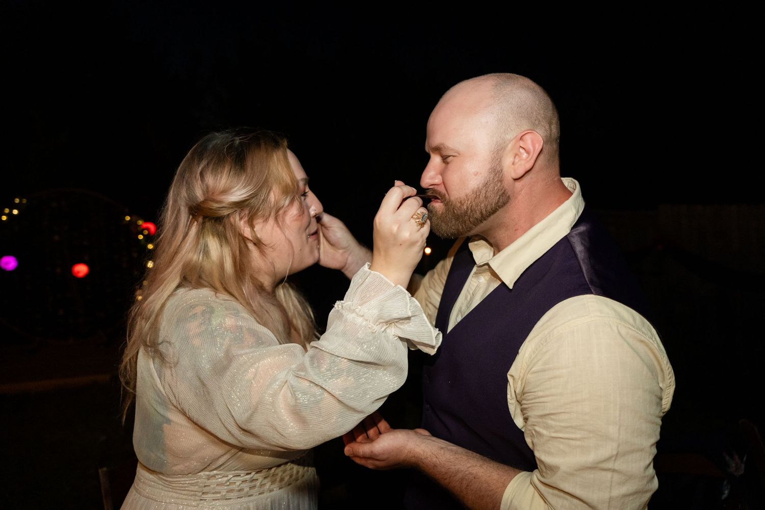 Couple feeds each other wedding cake at nighttime reception.