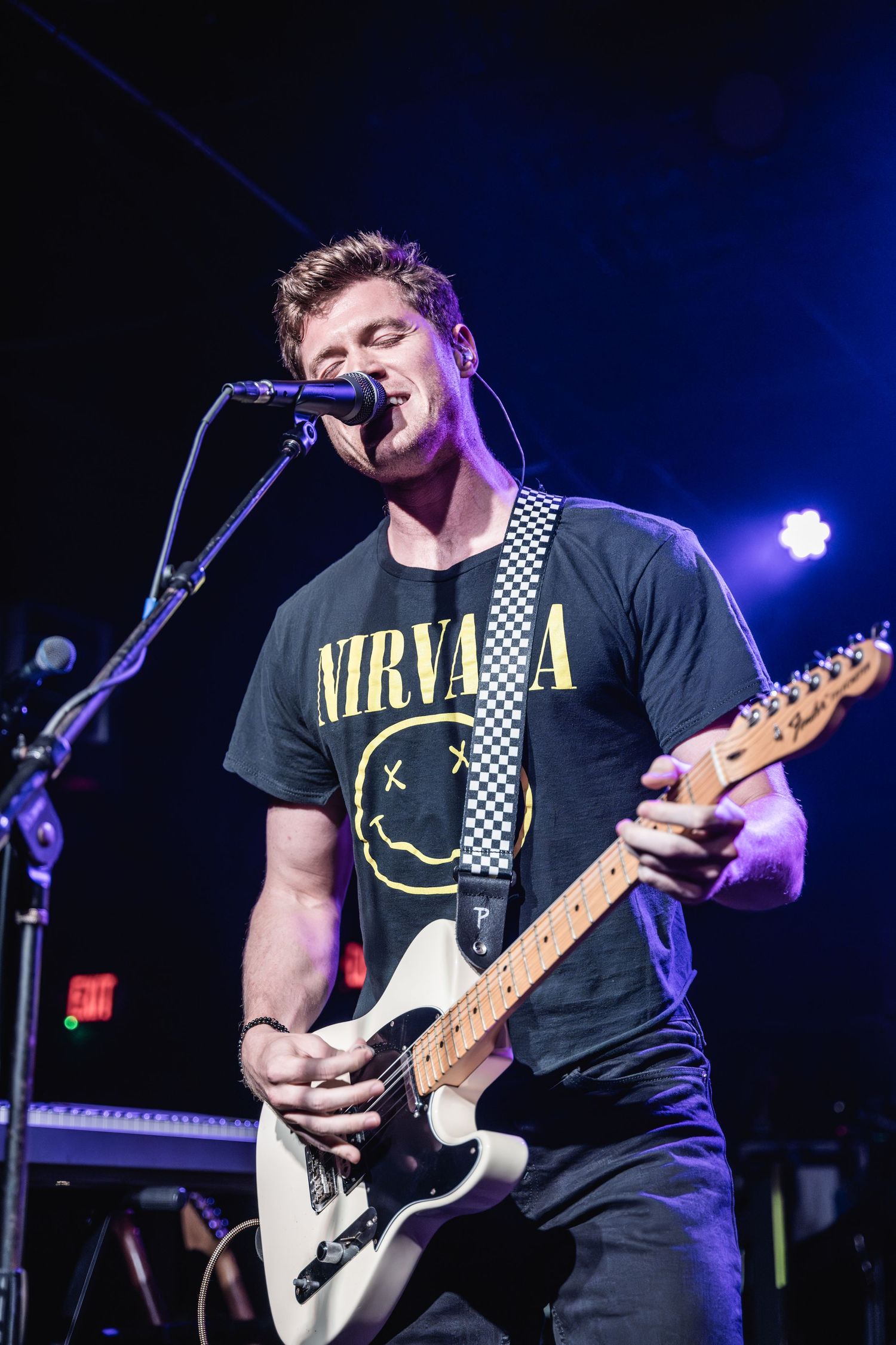 Musician performs on stage wearing Nirvana t-shirt while playing electric guitar and singing.