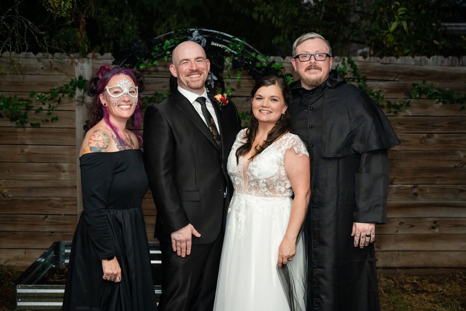 Small wedding party group poses against wooden fence backdrop in formal attire.