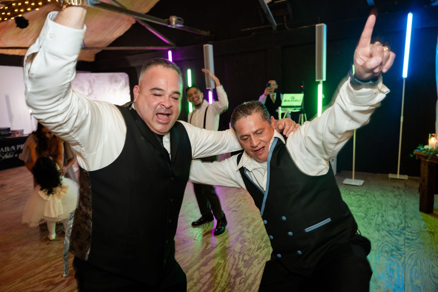 Two men in formal attire dancing enthusiastically with arms raised on a colorfully lit dance floor.