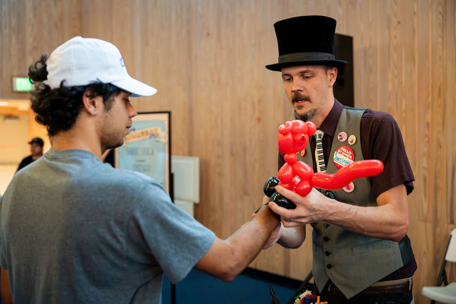 Two people interact as one wearing a top hat and vest demonstrates juggling with red pins at an indoor venue.
