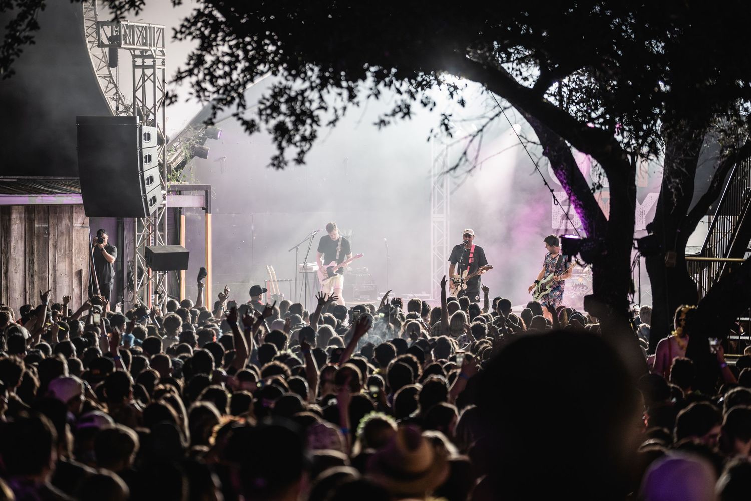Atmospheric black and white shot of festival crowd beneath trees with foggy stage lighting.