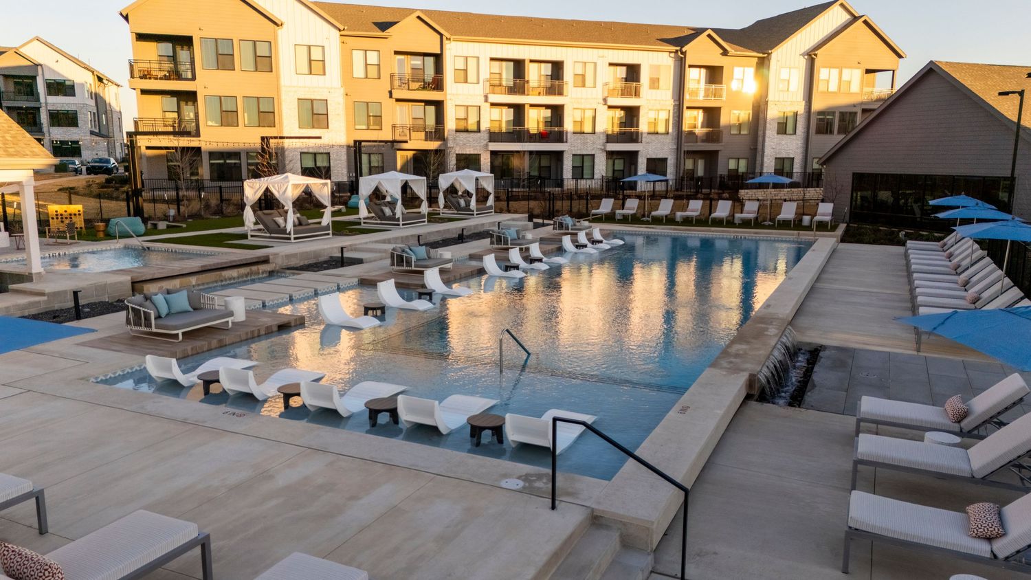 Modern apartment complex swimming pool illuminated at dusk with lounge chairs and cabanas around the perimeter.