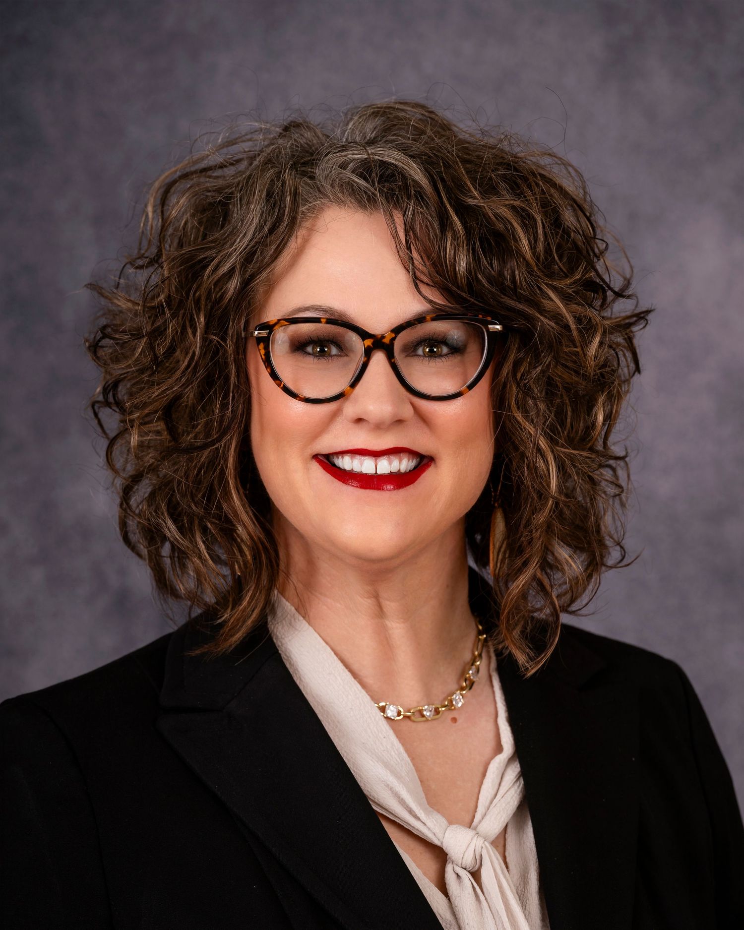 Professional headshot of a figure with curly brown hair, black cat-eye glasses, red lipstick and a black blazer with white blouse.