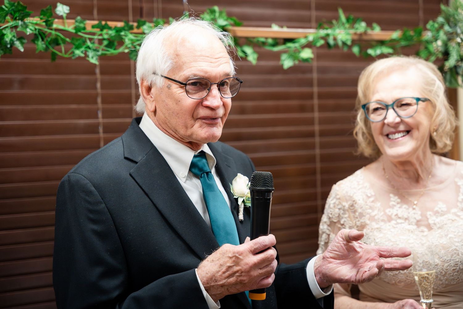 A well-dressed couple in formal attire share a joyful moment at a celebration with a wooden fence background.