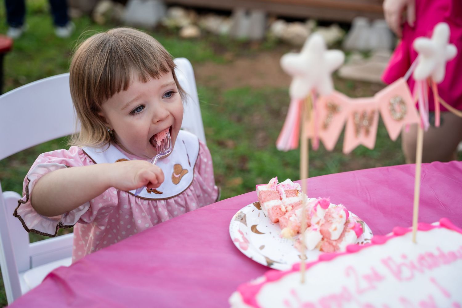A toddler in pink enjoys cake at a festive party with decorations.
