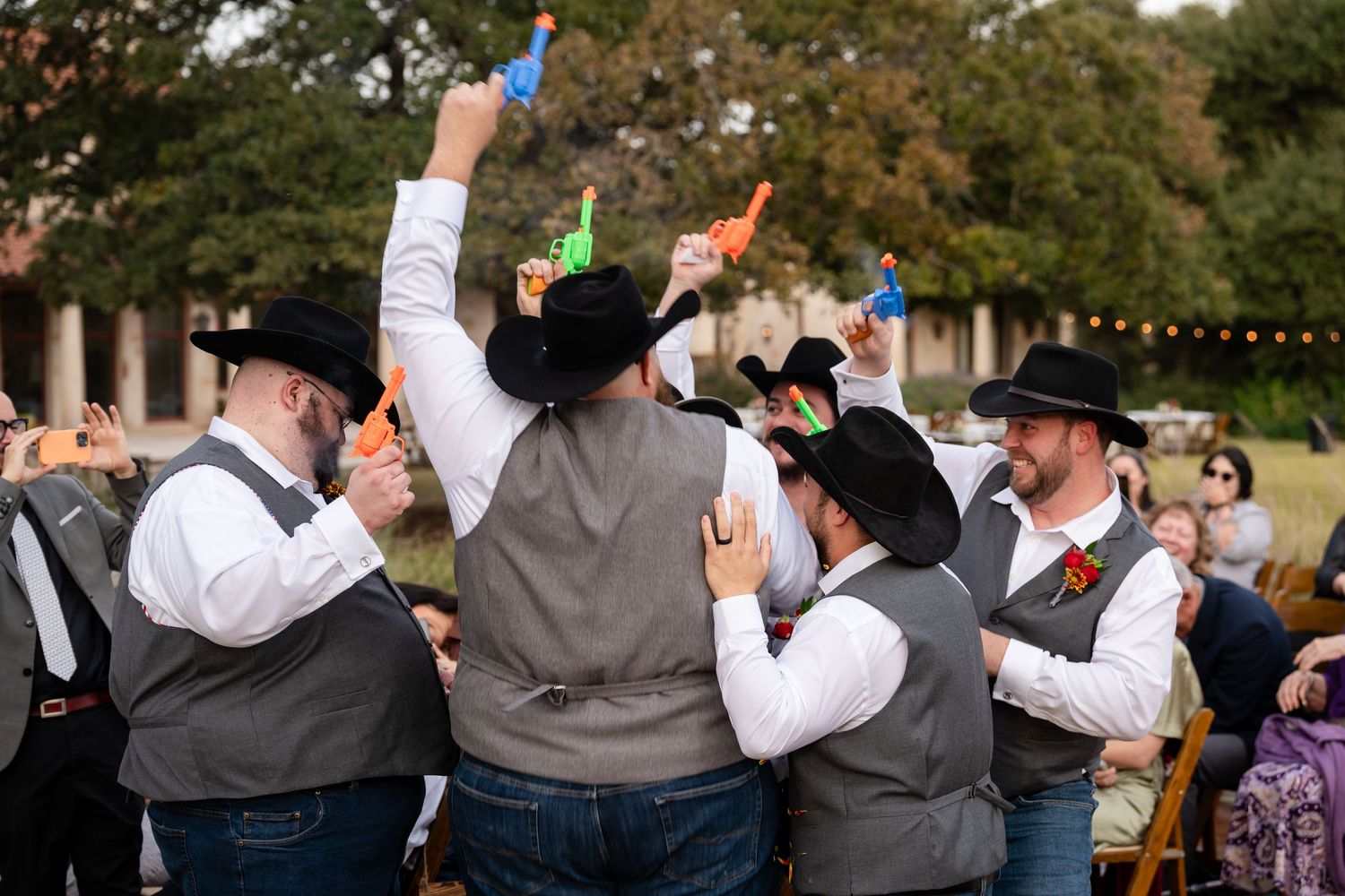 Groomsmen in cowboy hats and vests raise colorful drinks during outdoor wedding celebration.