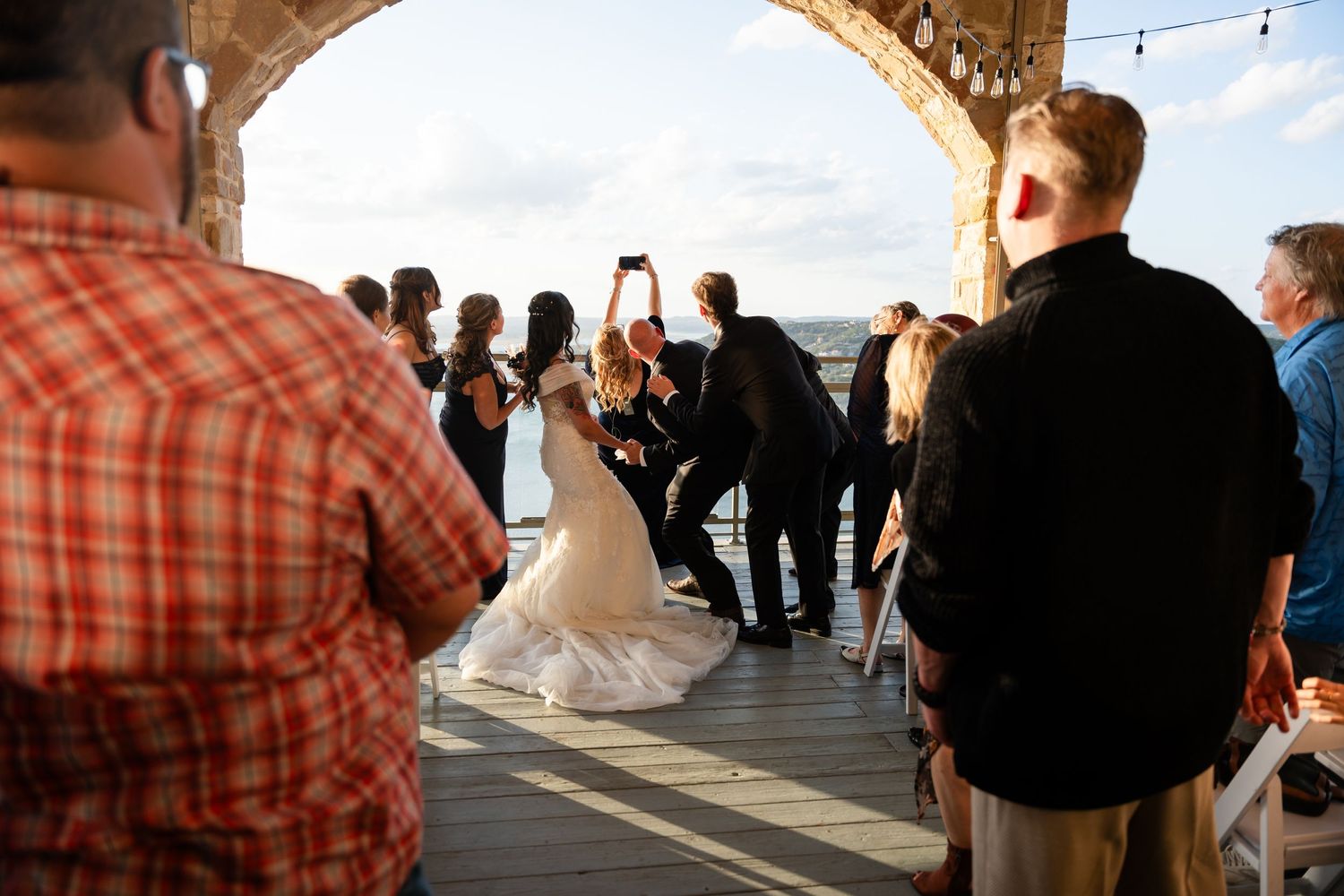 Wedding ceremony taking place under stone arch with ocean view and guests watching.