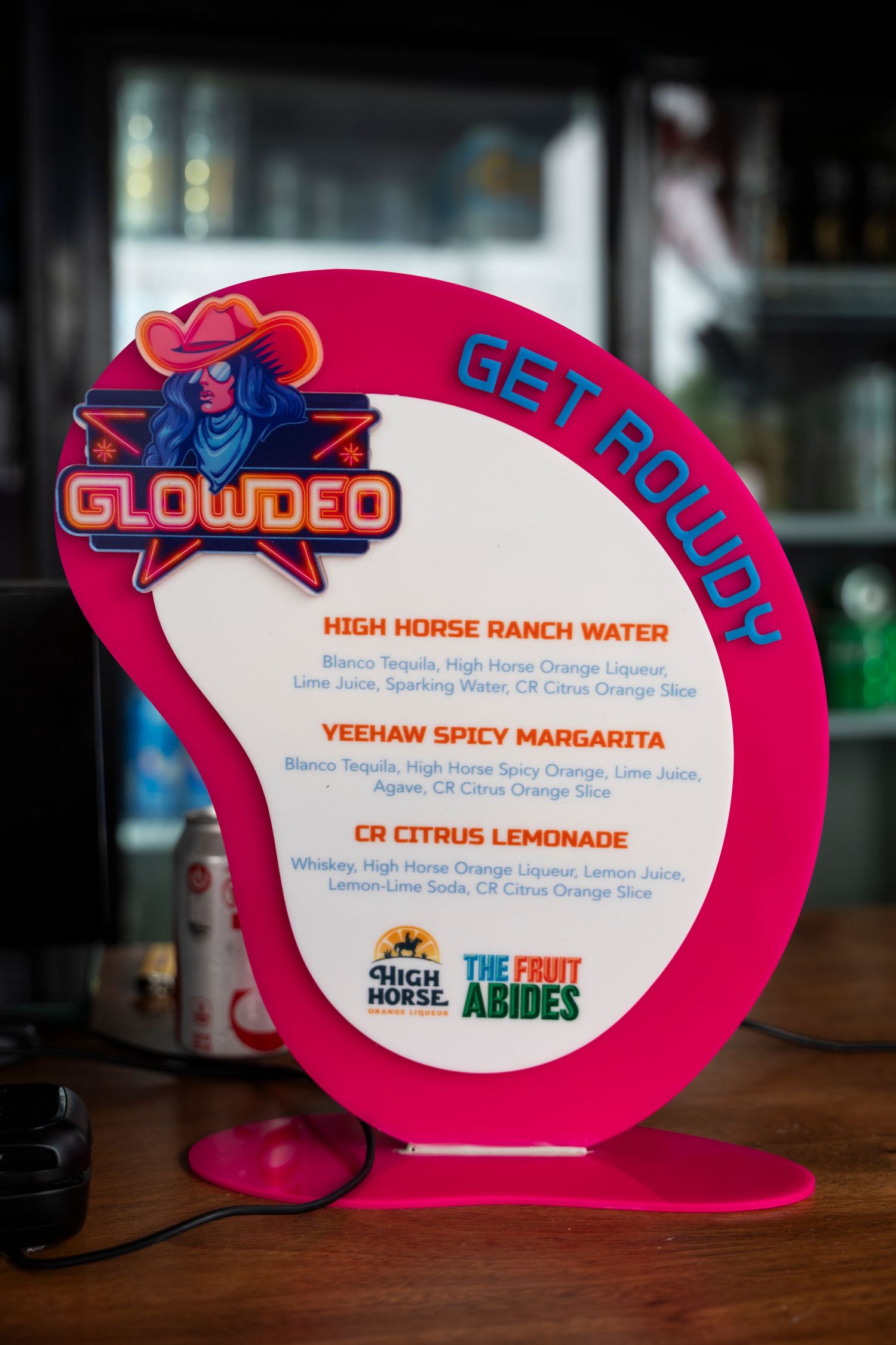 Illuminated pink neon menu signs displaying cocktail names and descriptions at a restaurant bar.