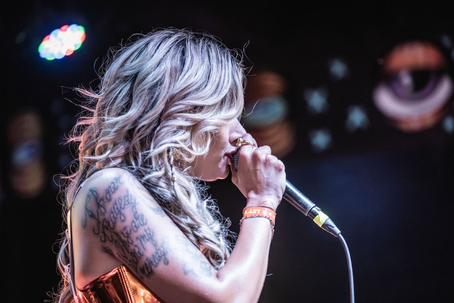 A vocalist with silver hair and colorful tattoos performs on stage with a microphone in dramatic lighting.