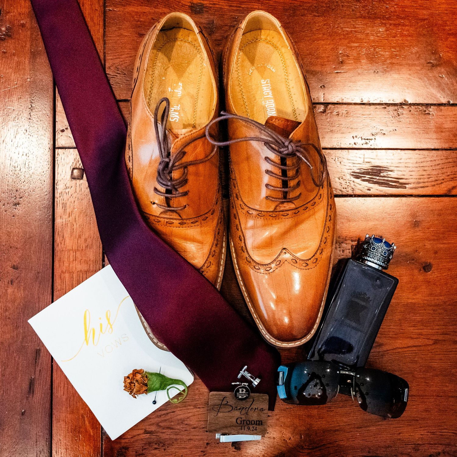 Brown leather dress shoes arranged with a burgundy tie and camera accessories on wooden flooring.
