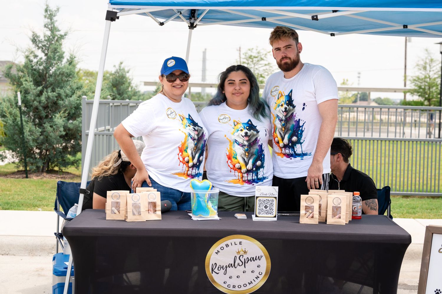 Event staff stand behind a merchandise table with branded items at an outdoor festival.
