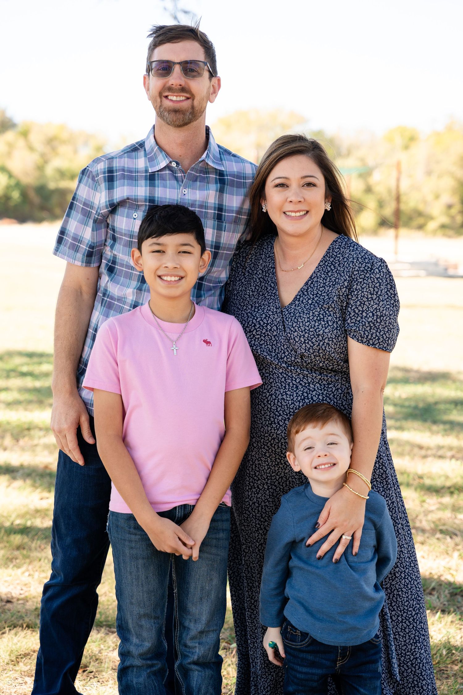 A happy family of four poses outdoors on a sunny day in casual clothing.