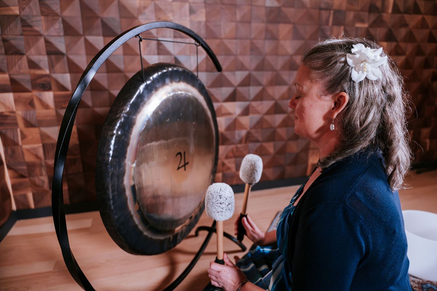 A person plays a large gong instrument against a geometric patterned wall.