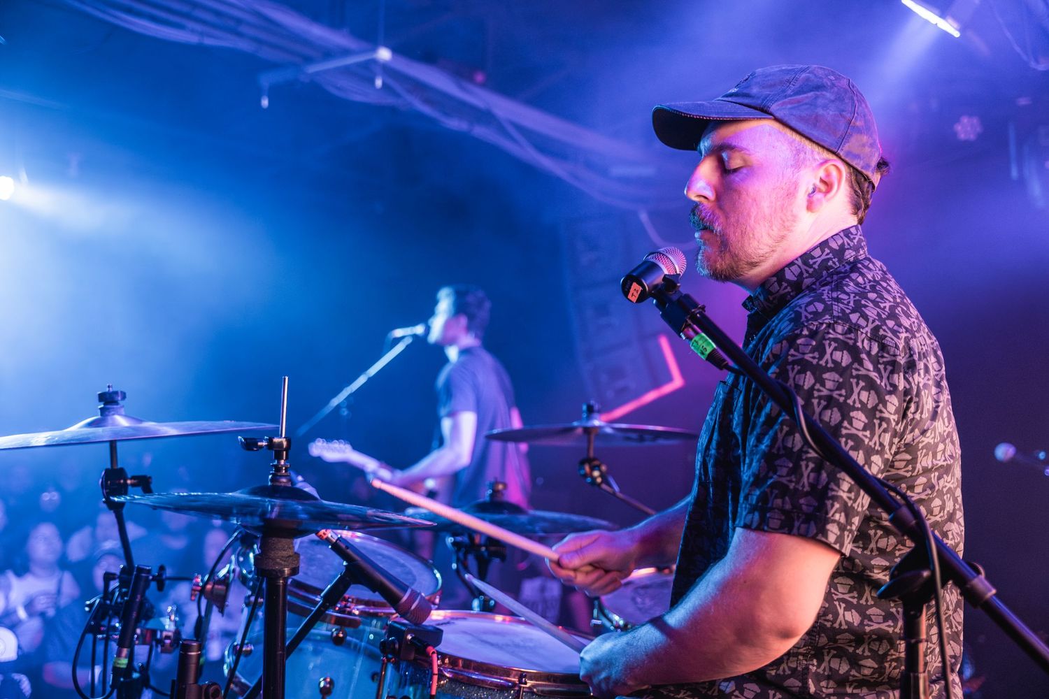 Musicians perform on stage with colorful blue lighting and drum kit visible in concert setting.
