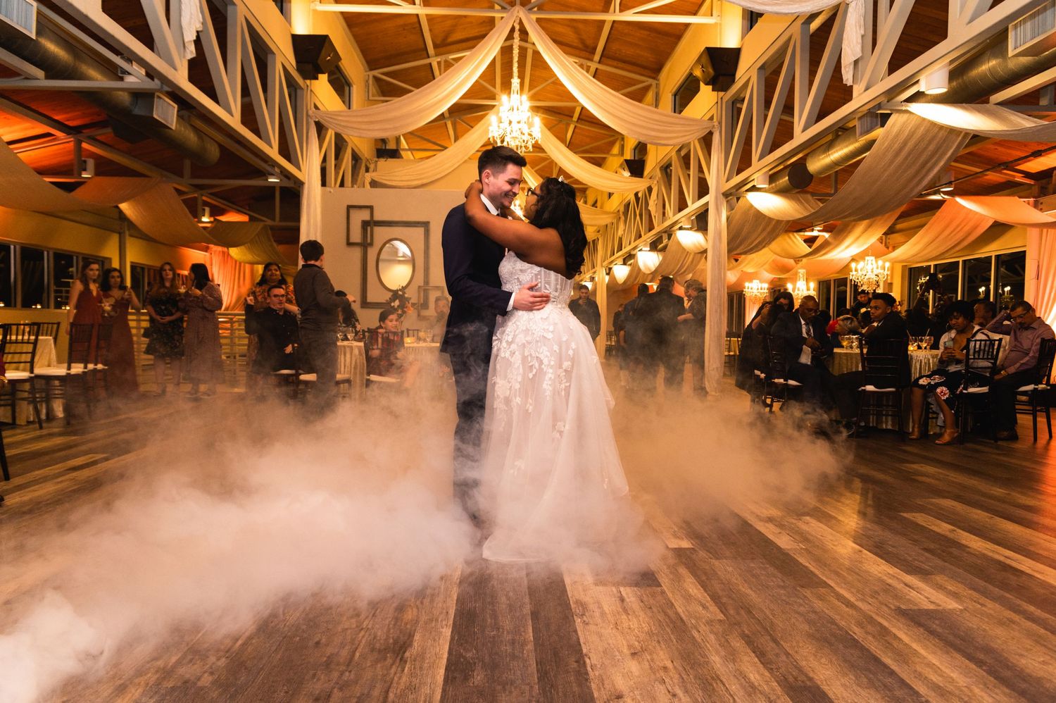 A dramatic fog-filled first dance on a wooden floor under decorative beams and chandeliers.