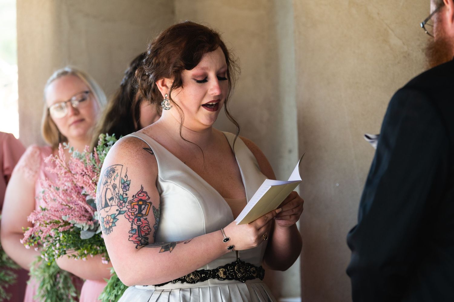 A wedding ceremony moment shows someone reading from a small book outdoors.
