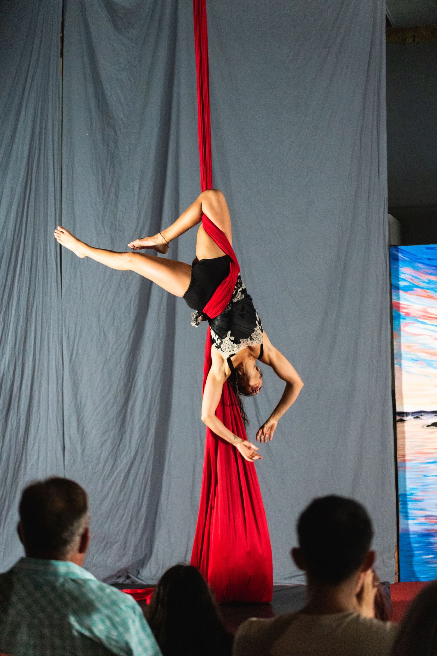 Aerialist performs inverted position on red silks before an audience against gray backdrop.