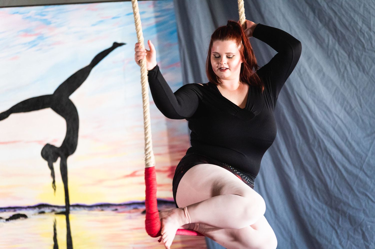 Performer in black attire poses on aerial rope with sunset mural backdrop.