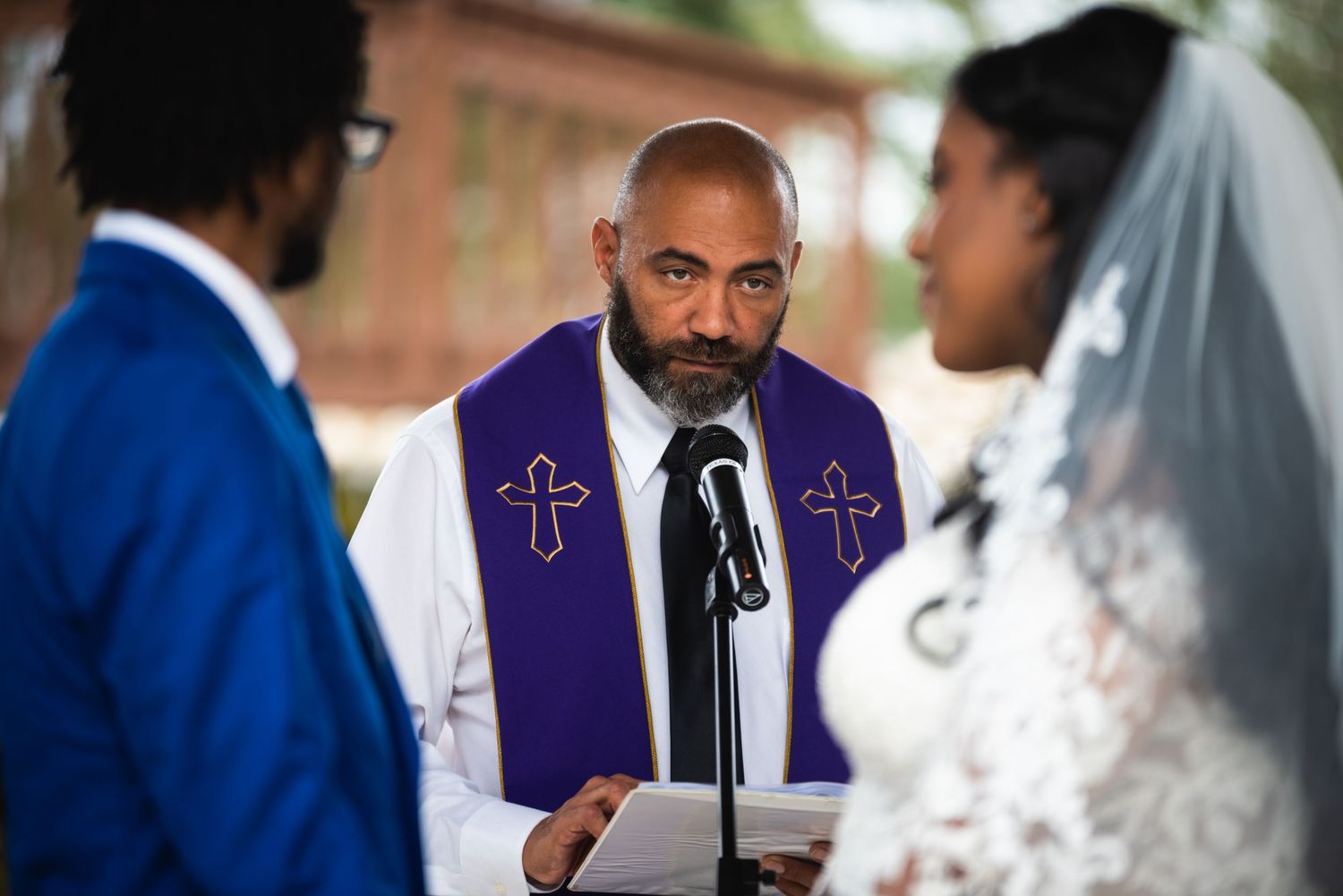 Minister in purple vestments leads outdoor wedding ceremony with cross embroidery detail.