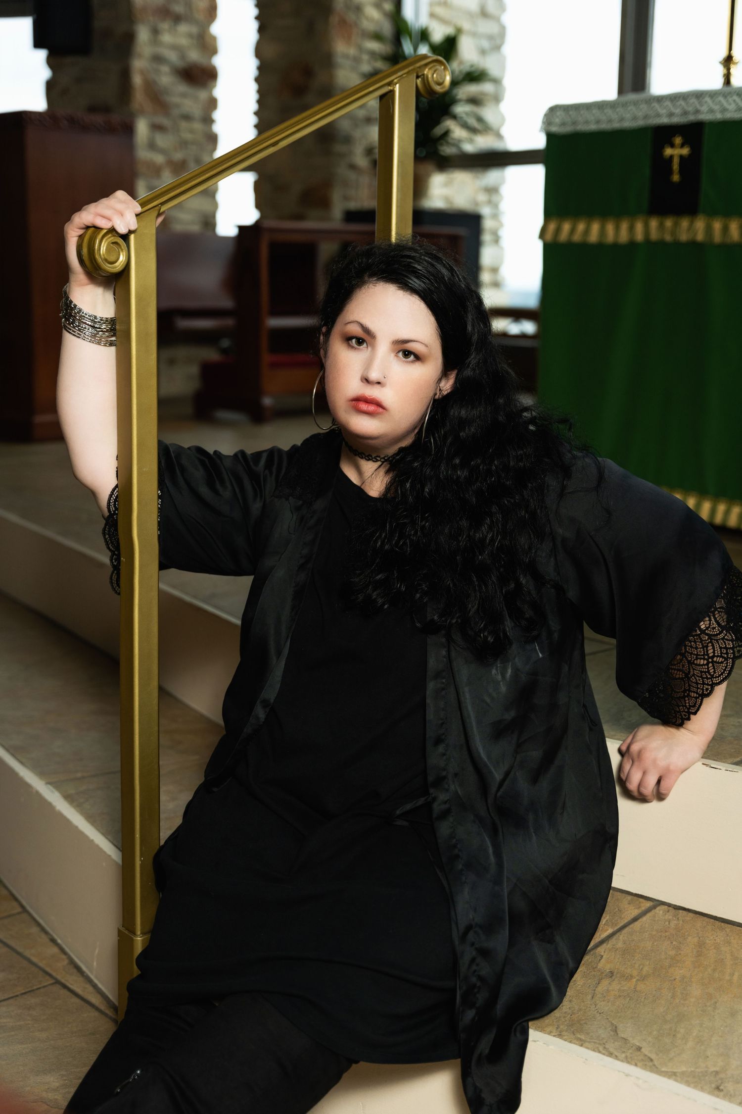 Someone in all black attire poses dramatically by a brass handrail in a church setting.