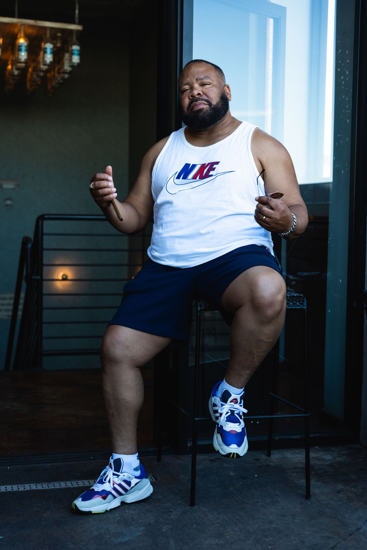 Person in white Nike tank top and navy shorts sitting on stool with expressive pose.
