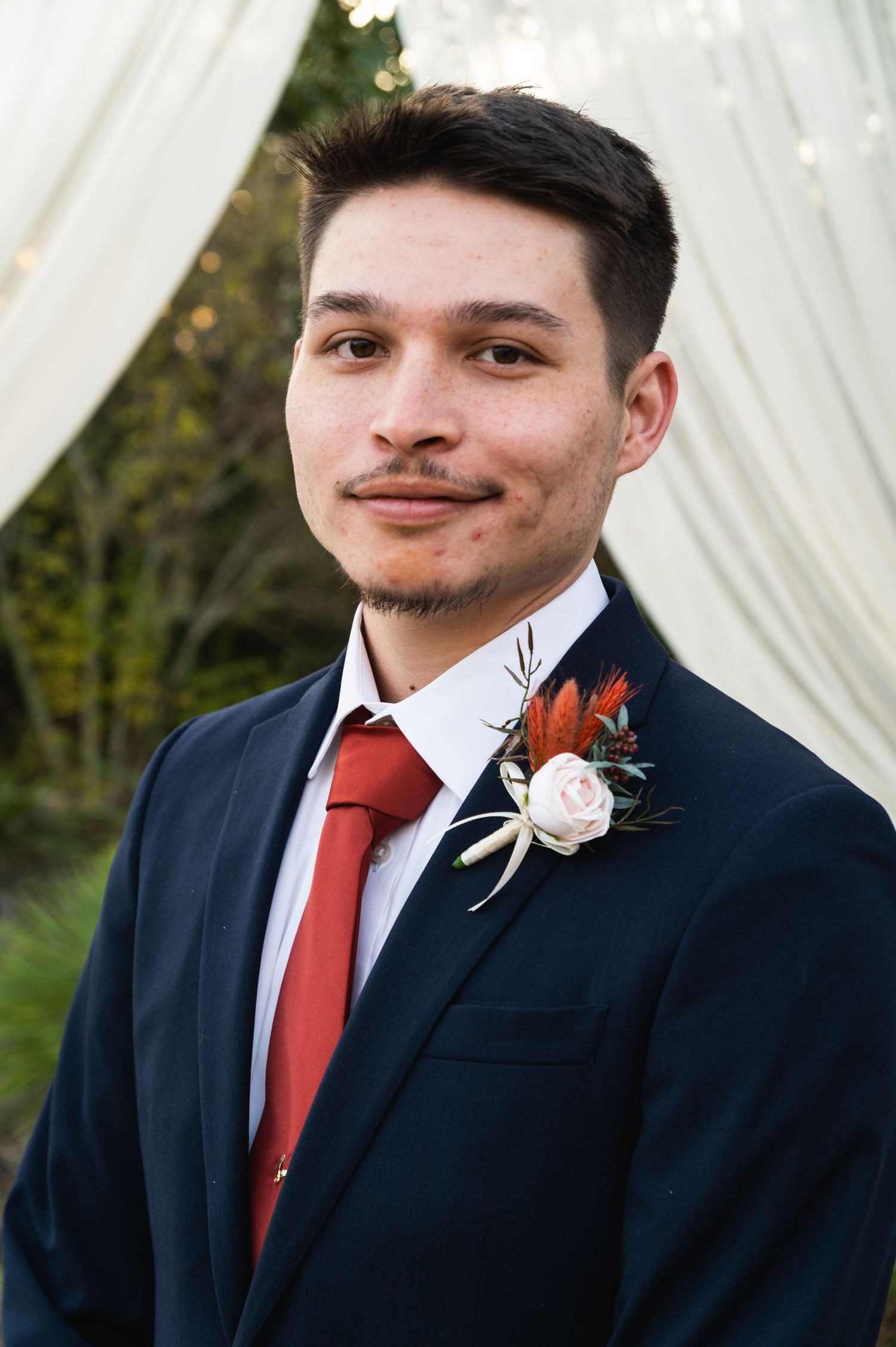 A groom in a navy suit with red tie and white boutonniere poses against white drapes.
