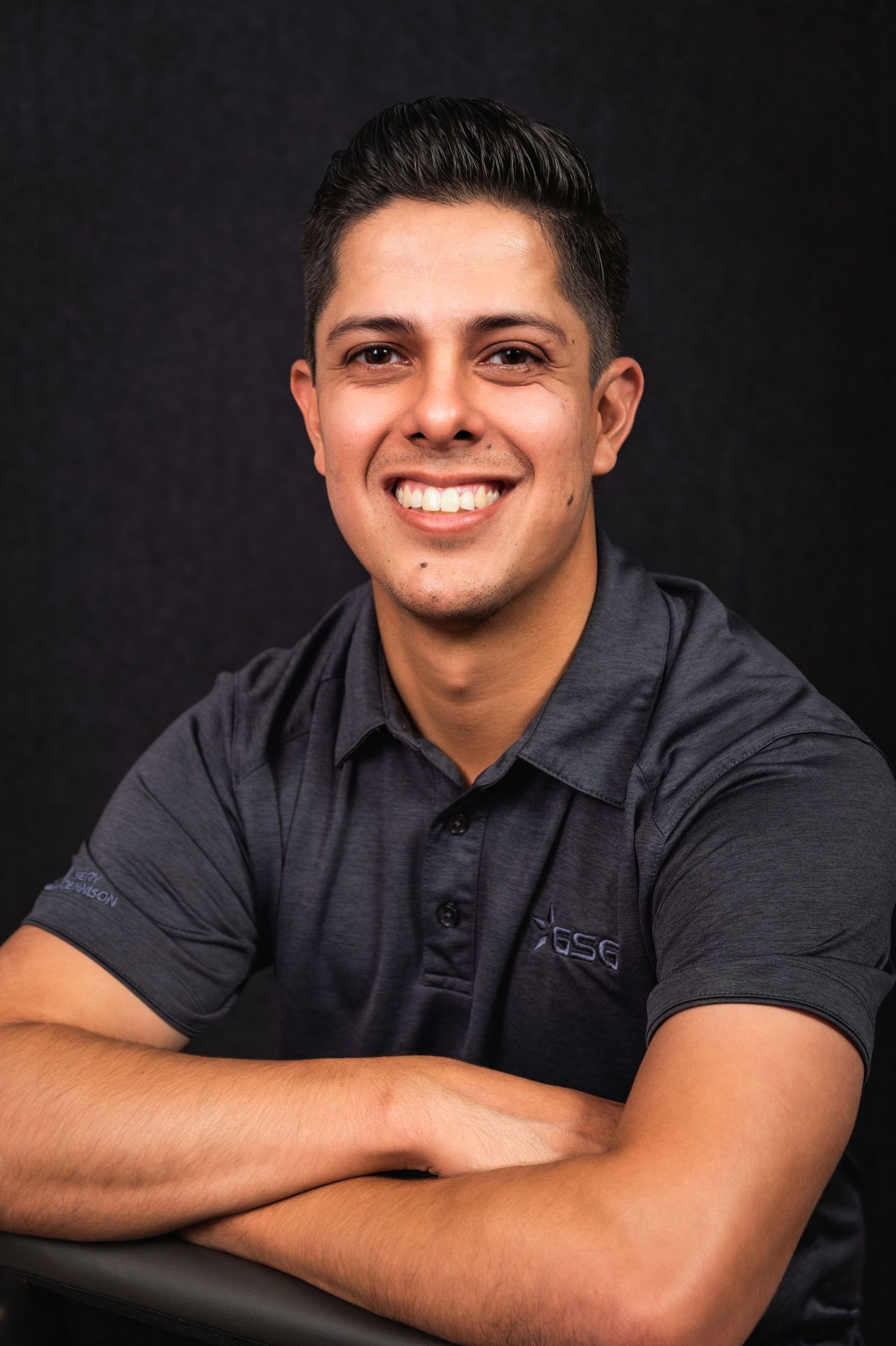 Professional headshot of person in dark polo shirt smiling against black background.