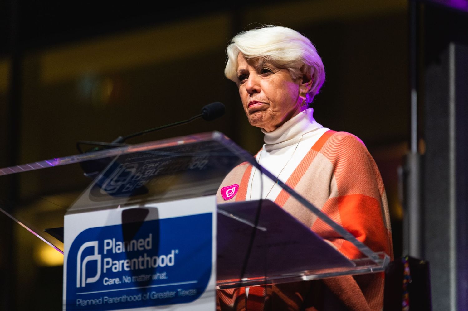 Speaker in red turtleneck addresses audience from clear acrylic podium with organizational logo.