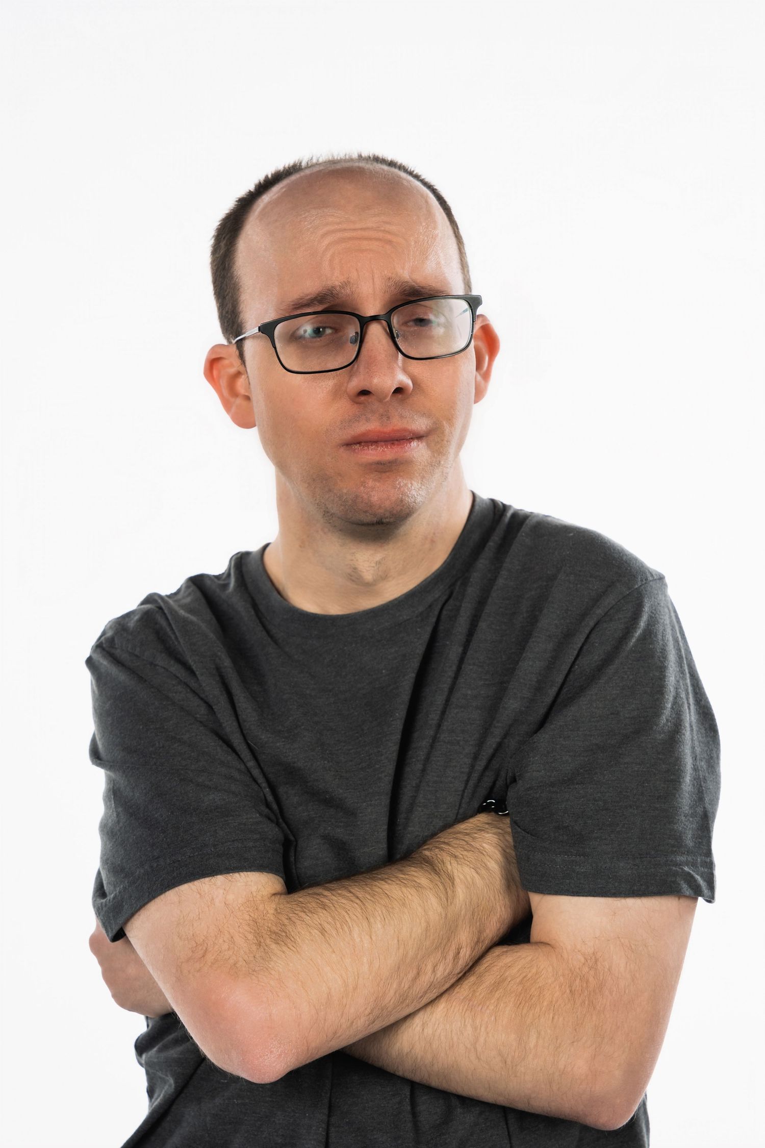A person in a grey t-shirt with glasses poses with arms crossed against a white background.