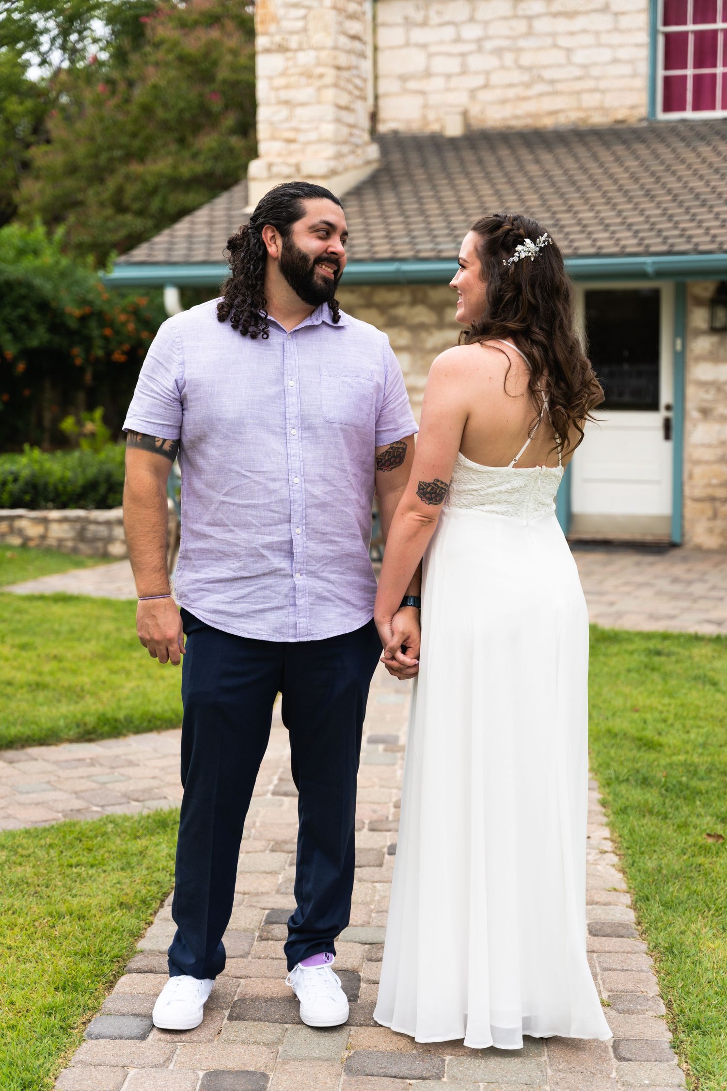 Couple in wedding attire standing on stone path in front of rustic building.