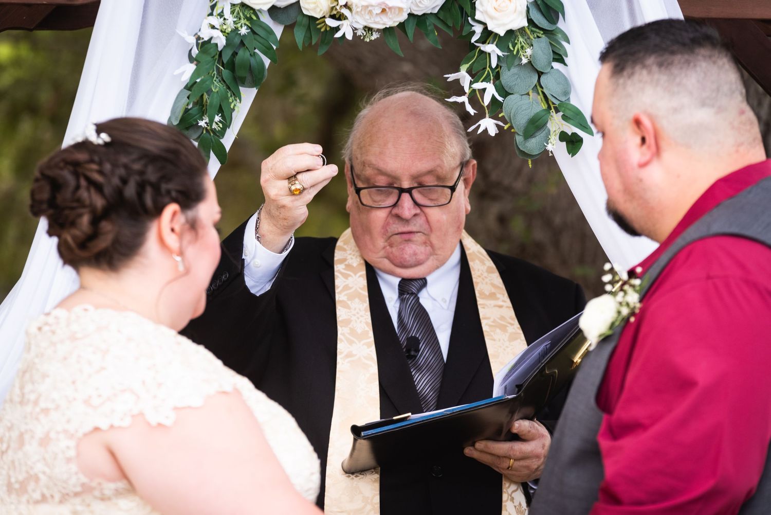 Wedding officiant holding book during outdoor ceremony beneath floral arch.