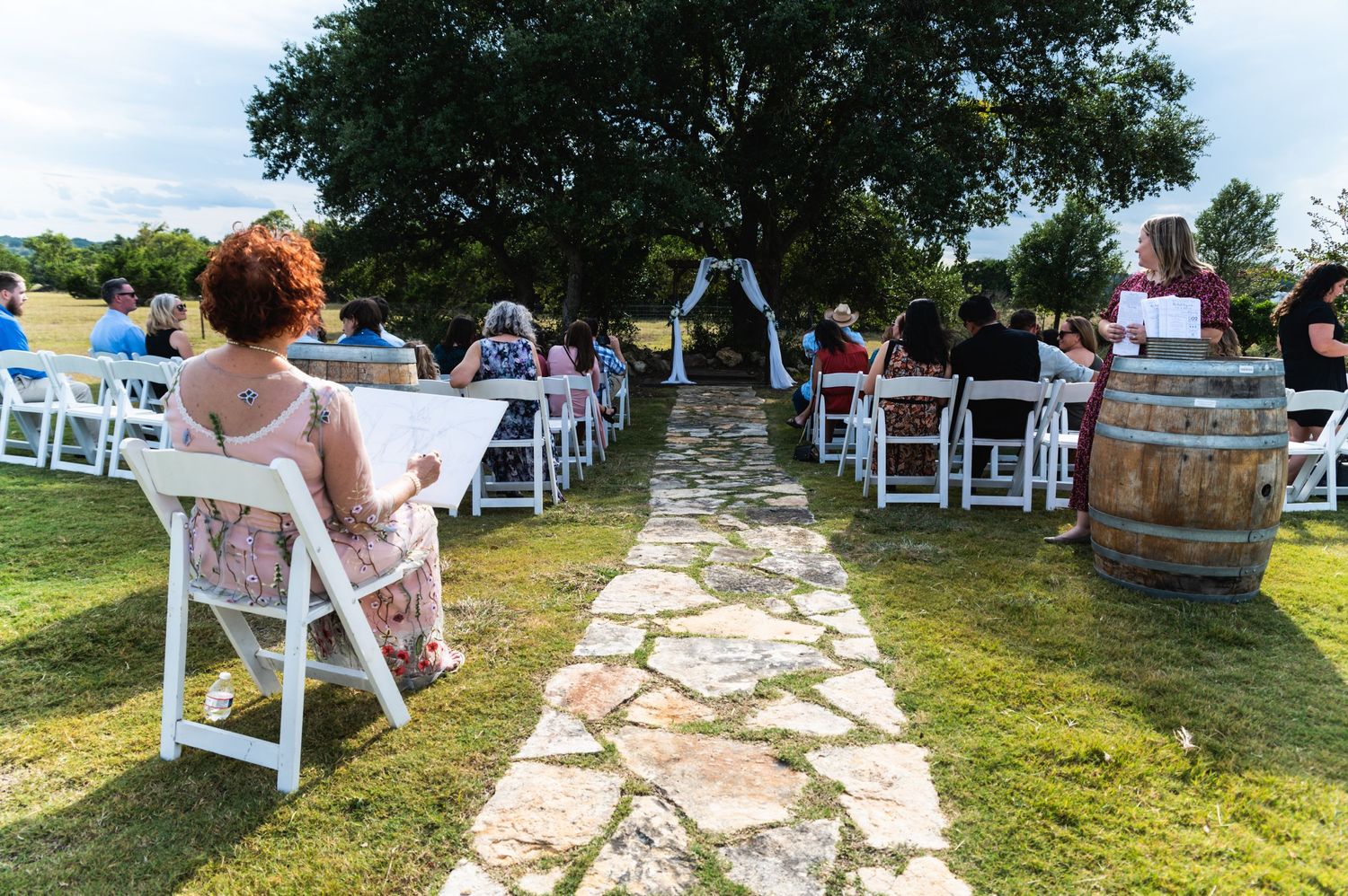 White chairs line a stone pathway at an outdoor wedding ceremony setting with wine barrels.