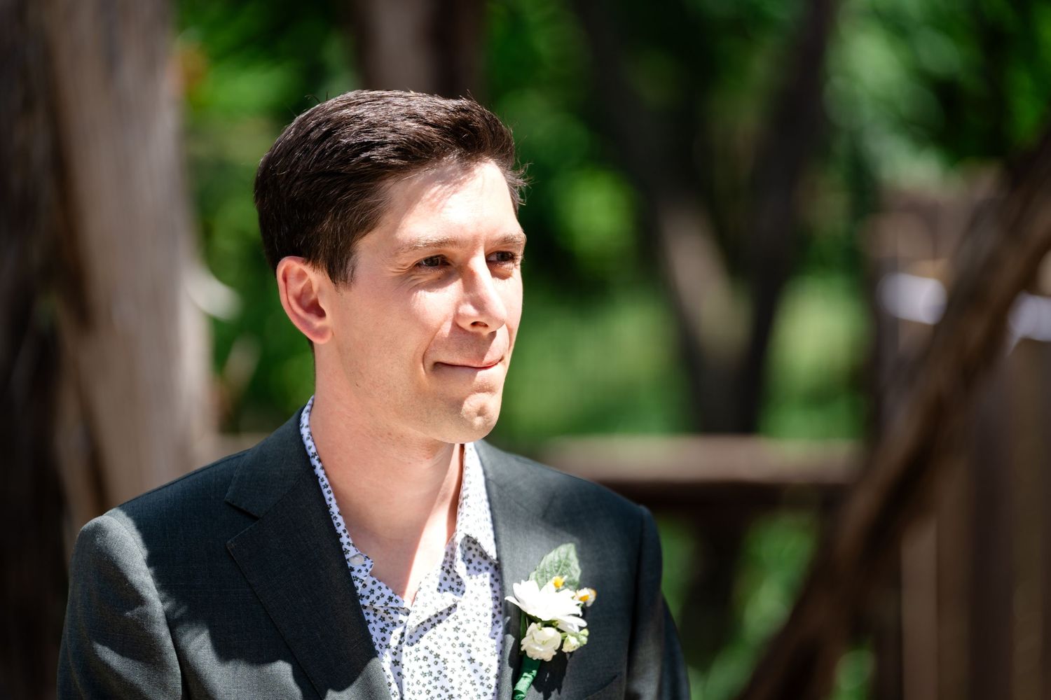 Groom in dark suit and patterned shirt smiles during outdoor wedding ceremony.